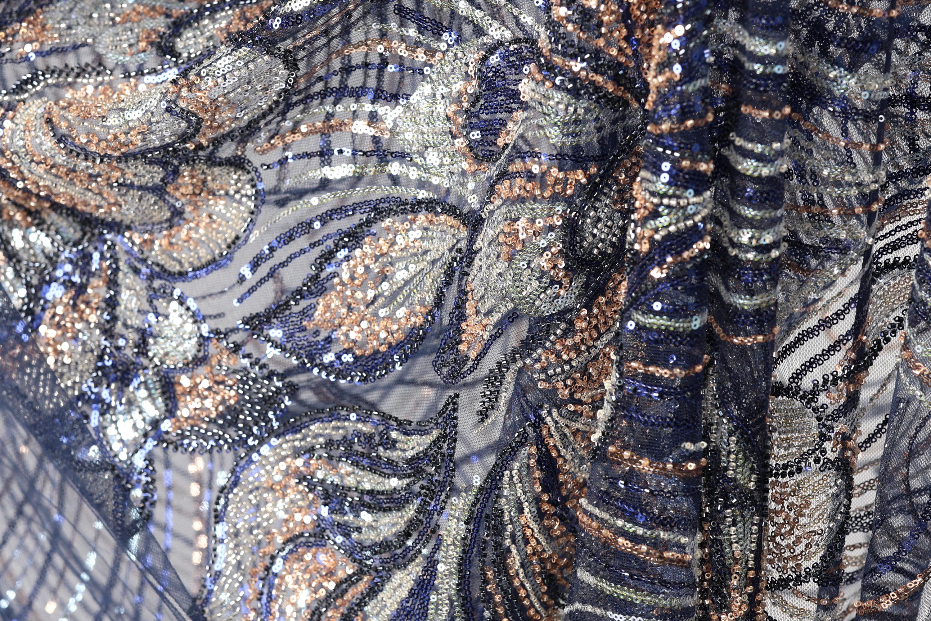 The image is a close-up of a fabric with a complex, intricate pattern of sequins and beads in shades of blue, gold, and silver. The pattern appears to be a floral or leaf-like design, with the sequins and beads arranged in a way that creates a visually striking and detailed appearance.