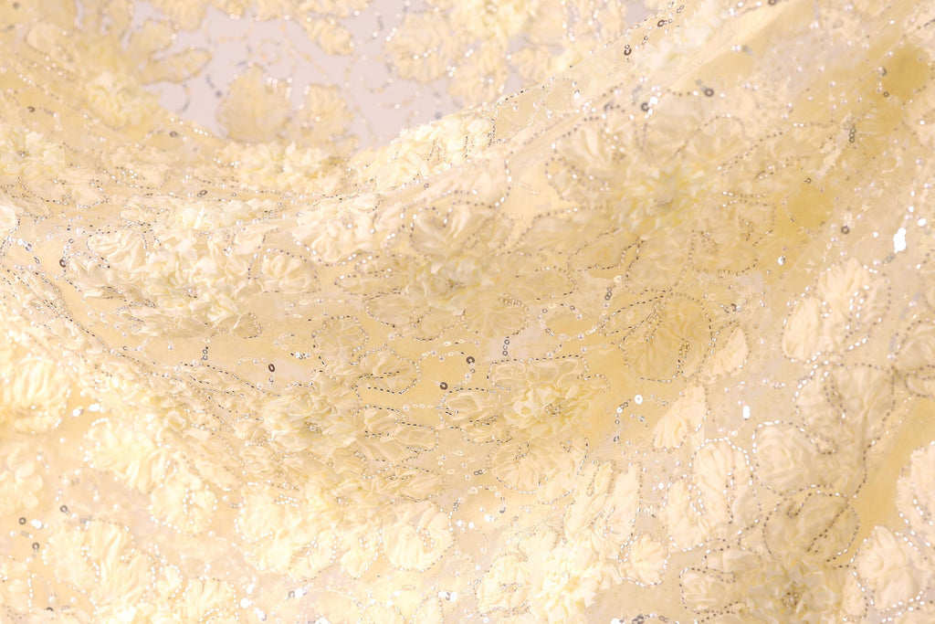 The image is a close-up of a fabric with a delicate, intricate floral pattern. The fabric appears to be made of a soft, light-colored material, possibly silk or chiffon, with a floral design that includes white flowers and leaves. The fabric has a shimmering, sparkly effect, adding a touch of elegance and sophistication to the overall appearance.