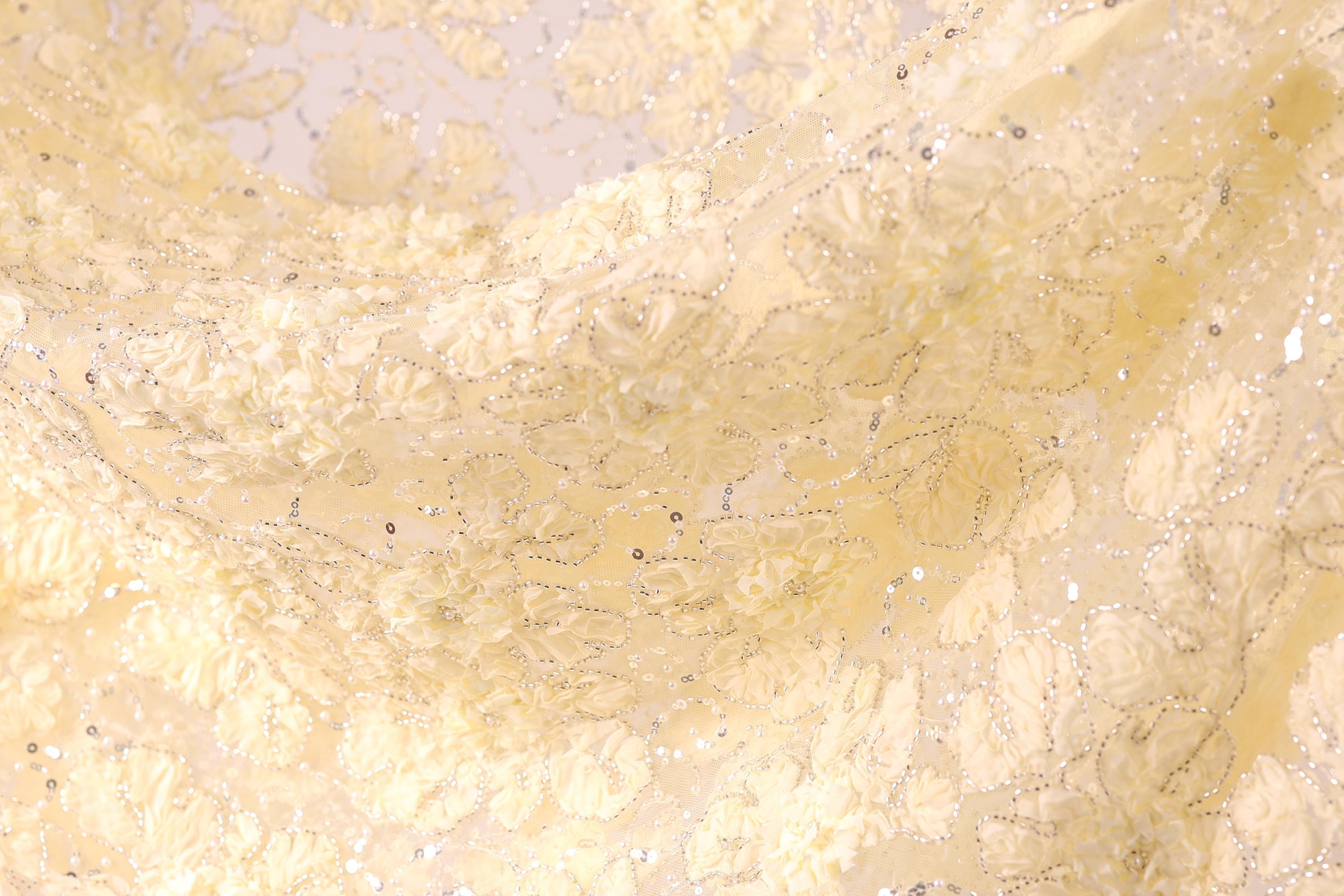 The image is a close-up of a fabric with a delicate, intricate floral pattern. The fabric appears to be made of a soft, light-colored material, possibly silk or chiffon, with a floral design that includes white flowers and leaves. The fabric has a shimmering, sparkly effect, adding a touch of elegance and sophistication to the overall appearance.