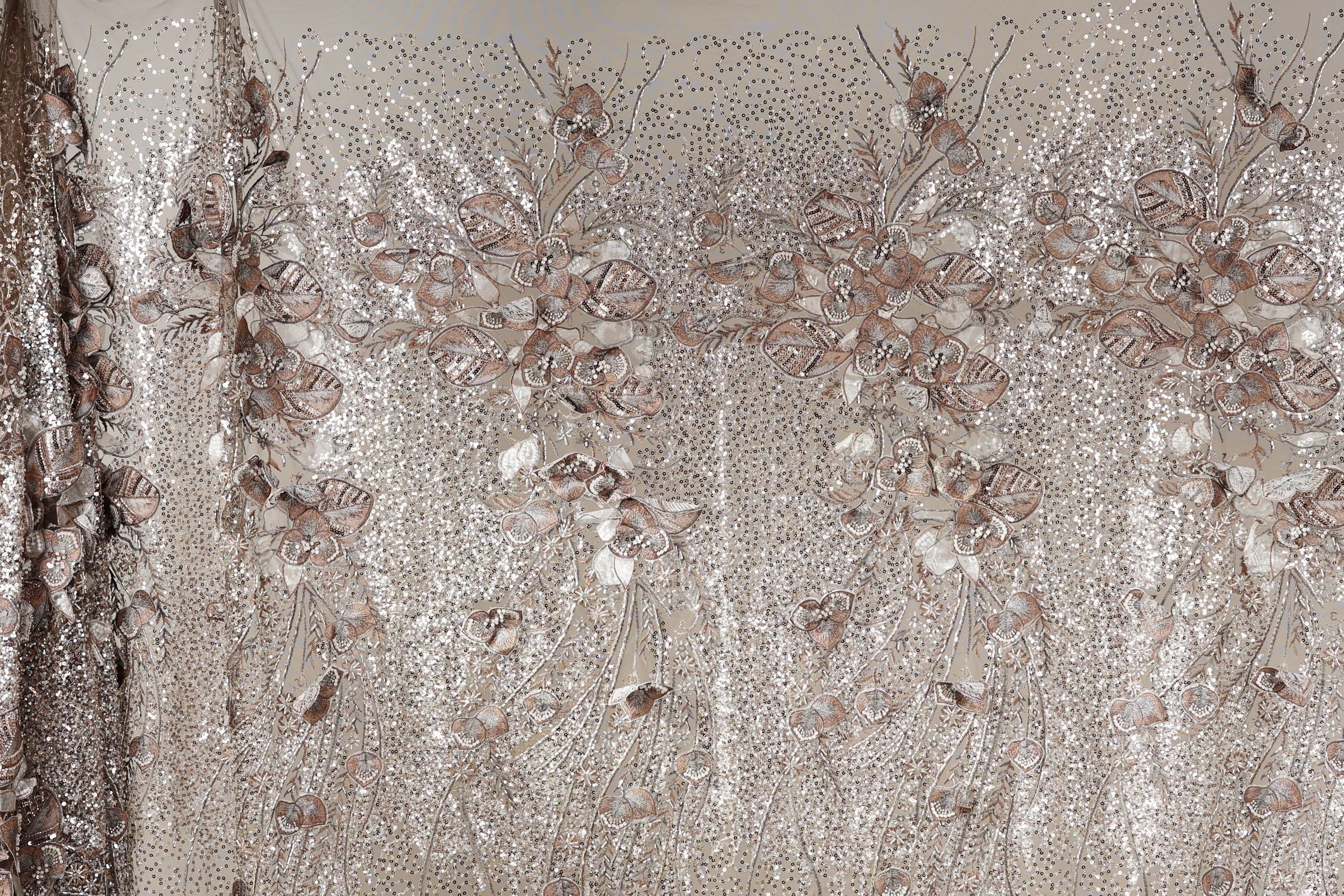 a close-up view of a fabric with a floral pattern, including various shades of brown and beige. The fabric appears to be made of sequin material, giving it a shiny and decorative appearance.