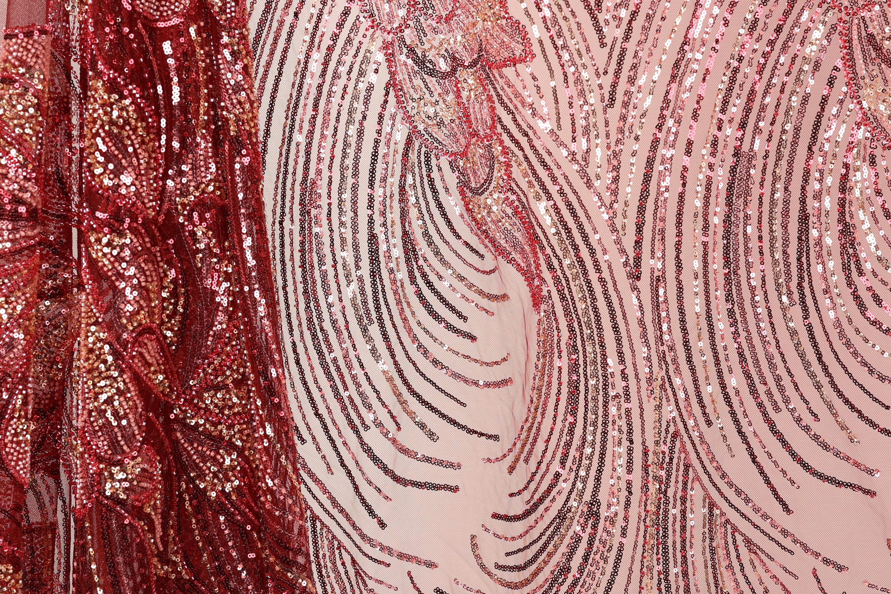a close-up view of a fabric with a striking pattern of red and pink sequins. The fabric appears to be a sheer material, with the sequins arranged in a swirling, wave-like design. The background of the image is a light pink color, which complements the vibrant colors of the sequins. The image does not contain any discernible text or countable objects, and the focus is solely on the intricate and visually appealing pattern of the sequins.