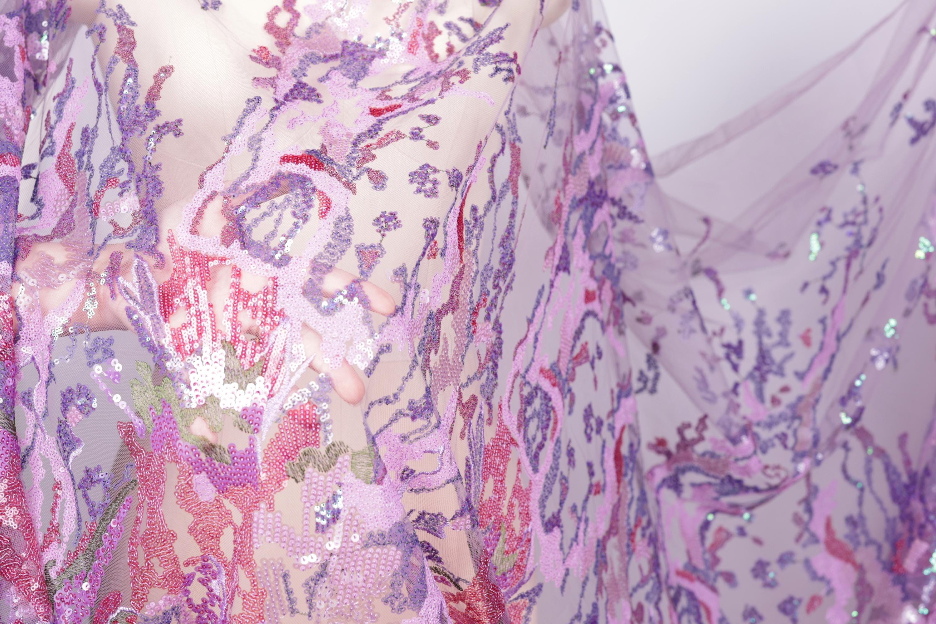 The image is a close-up view of a fabric with a floral pattern, featuring a mix of pink, purple, and green colors. The fabric appears to be made of sequins or other embellishments, adding a sparkly and decorative touch.