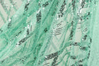 a close-up view of a fabric with a green and black sequin pattern. The fabric appears to be made of sheer material, with a metallic sheen due to the sequins. The pattern is intricate and detailed, with the sequins arranged in a way that creates a visually striking effect.