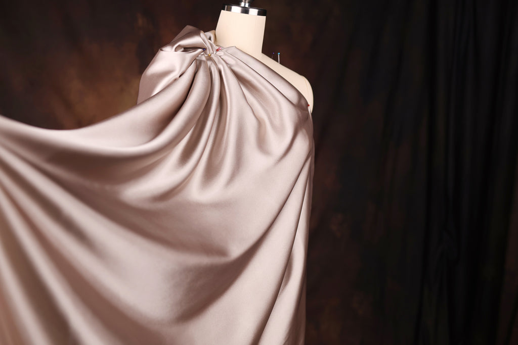 A mannequin wearing a flowing, draped fabric in a light pink color, set against a dark background.