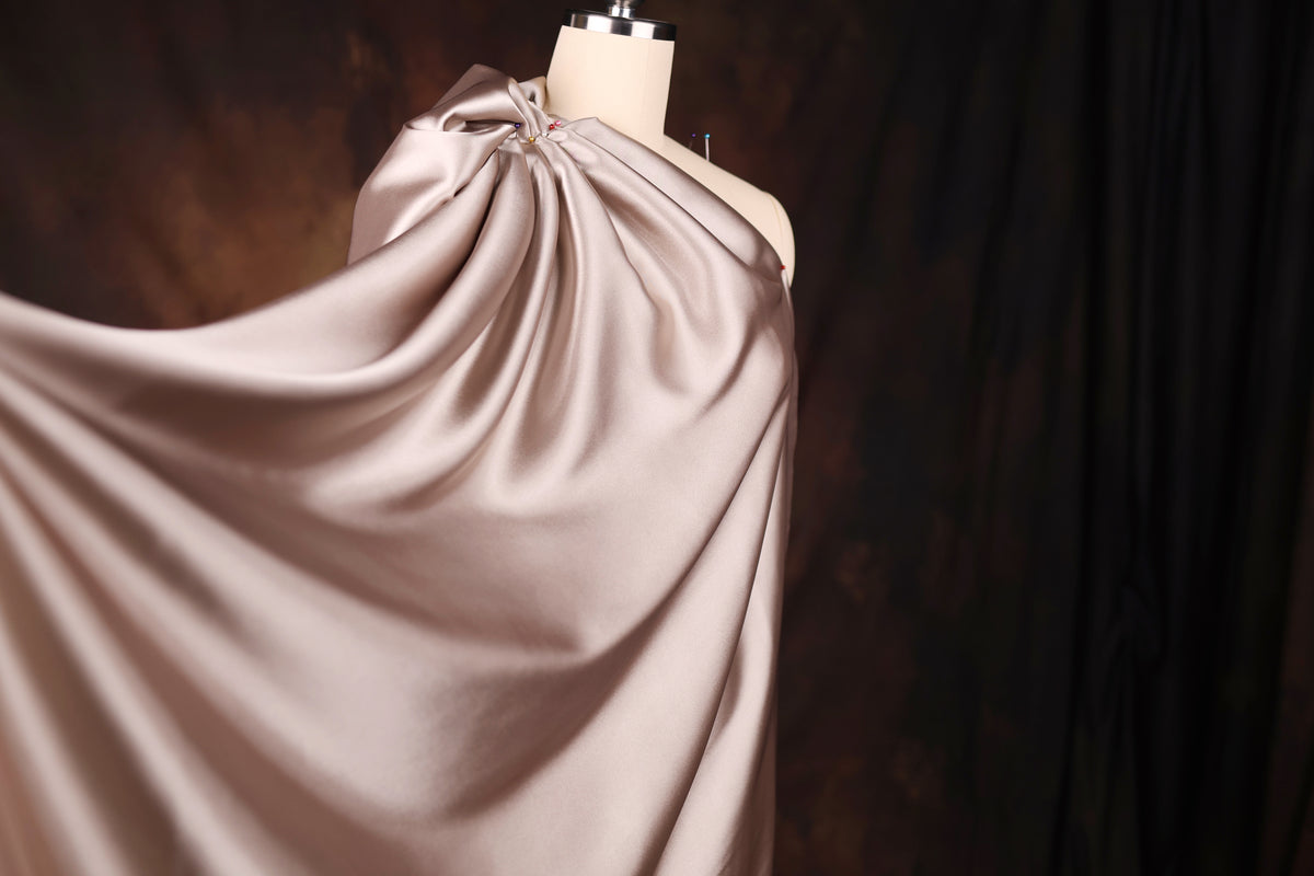 A mannequin wearing a flowing, draped fabric in a light pink color, set against a dark background.