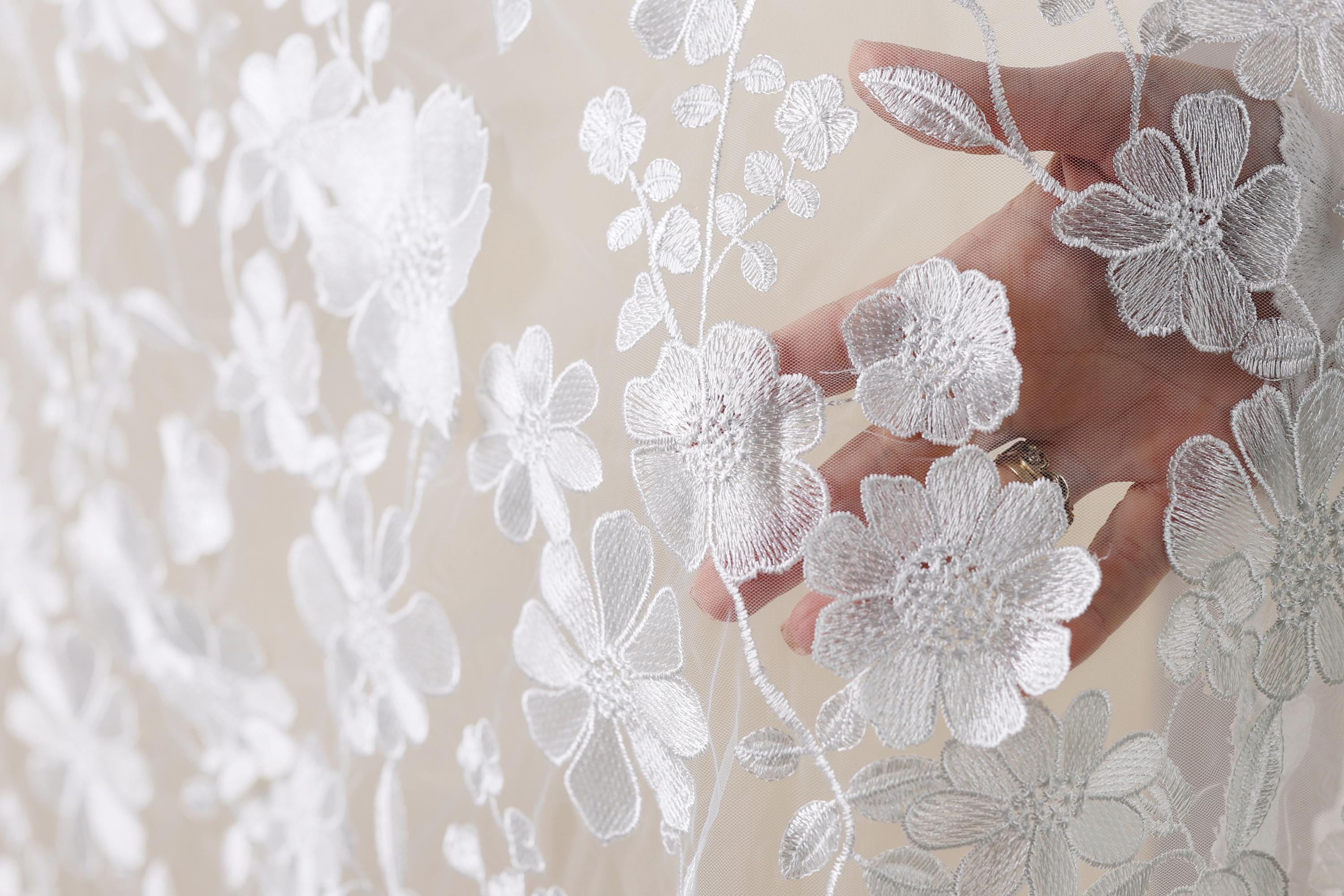 A hand is holding a delicate white lace fabric with intricate floral embroidery.