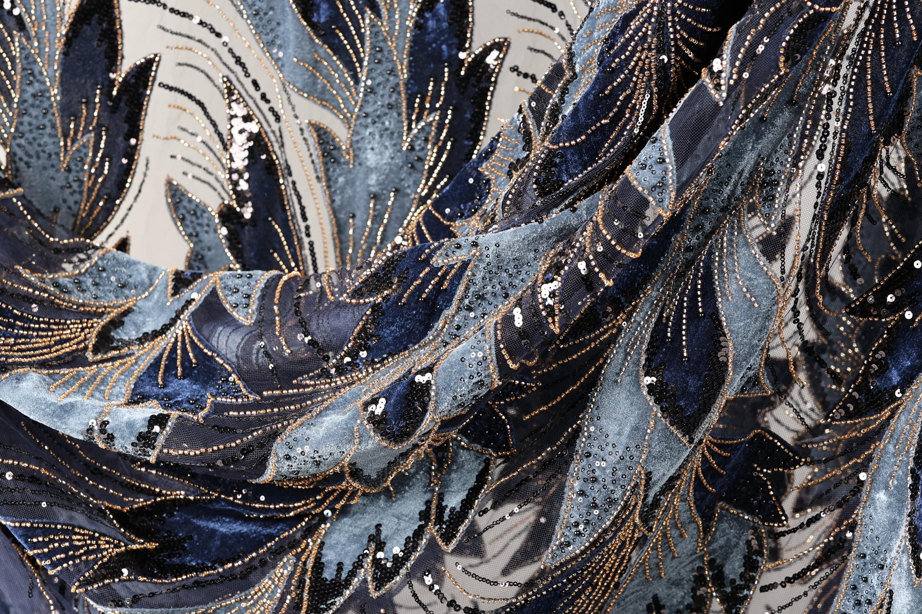 The image is a close-up view of a fabric with a complex, intricate pattern of blue and black sequins and beads. The pattern appears to be a mix of different textures and colors, creating a visually striking and detailed design.