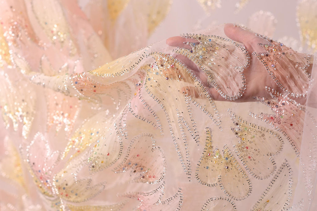 A close-up view of a delicate, hand-embroidered fabric with a floral pattern. The fabric appears to be made of sheer material, with a light pink and white color scheme. The intricate embroidery and beading on the fabric create a visually appealing and delicate appearance.