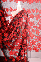 a close-up view of a fabric with a striking red floral pattern. The fabric appears to be a sheer material, with a black and red color scheme. The pattern consists of numerous red flowers arranged in a repeating design, creating a visually captivating and vibrant appearance.