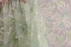 a close-up view of a delicate, intricately embroidered fabric with a floral pattern. The fabric appears to be made of a light green or mint-colored material, adorned with small, multicolored beads or sequins that add a touch of sparkle and detail to the design. The background of the image is blurred, drawing focus to the fabric and its details.