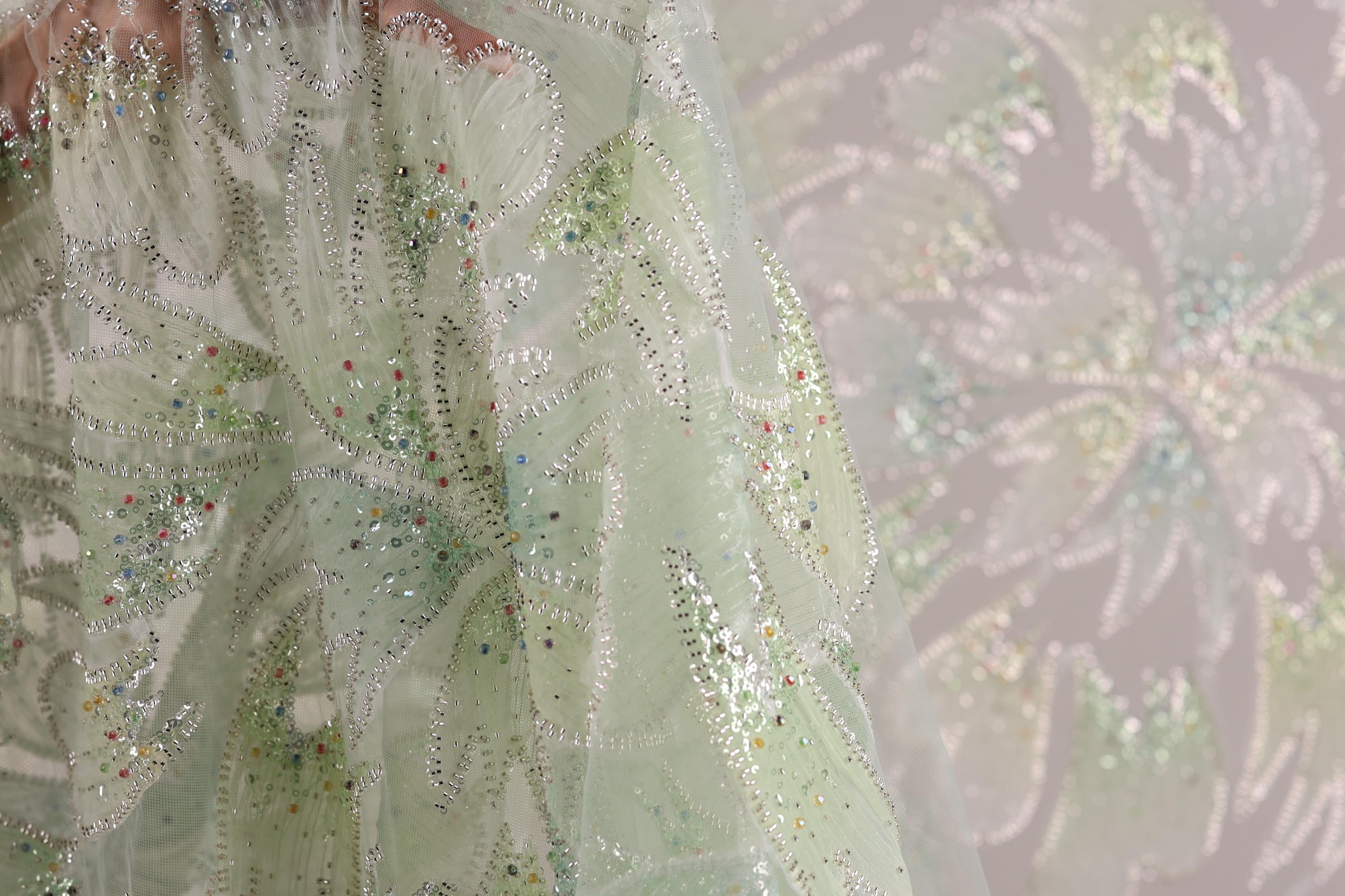 a close-up view of a delicate, intricately embroidered fabric with a floral pattern. The fabric appears to be made of a light green or mint-colored material, adorned with small, multicolored beads or sequins that add a touch of sparkle and detail to the design. The background of the image is blurred, drawing focus to the fabric and its details.