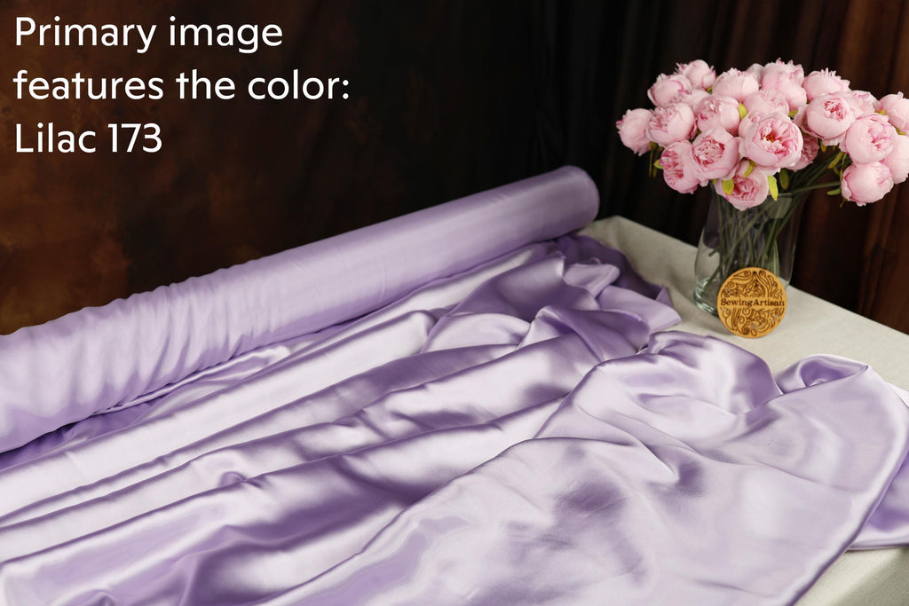 a roll of purple satin fabric, a vase of pink peonies, and a gold medal, all arranged on a table.