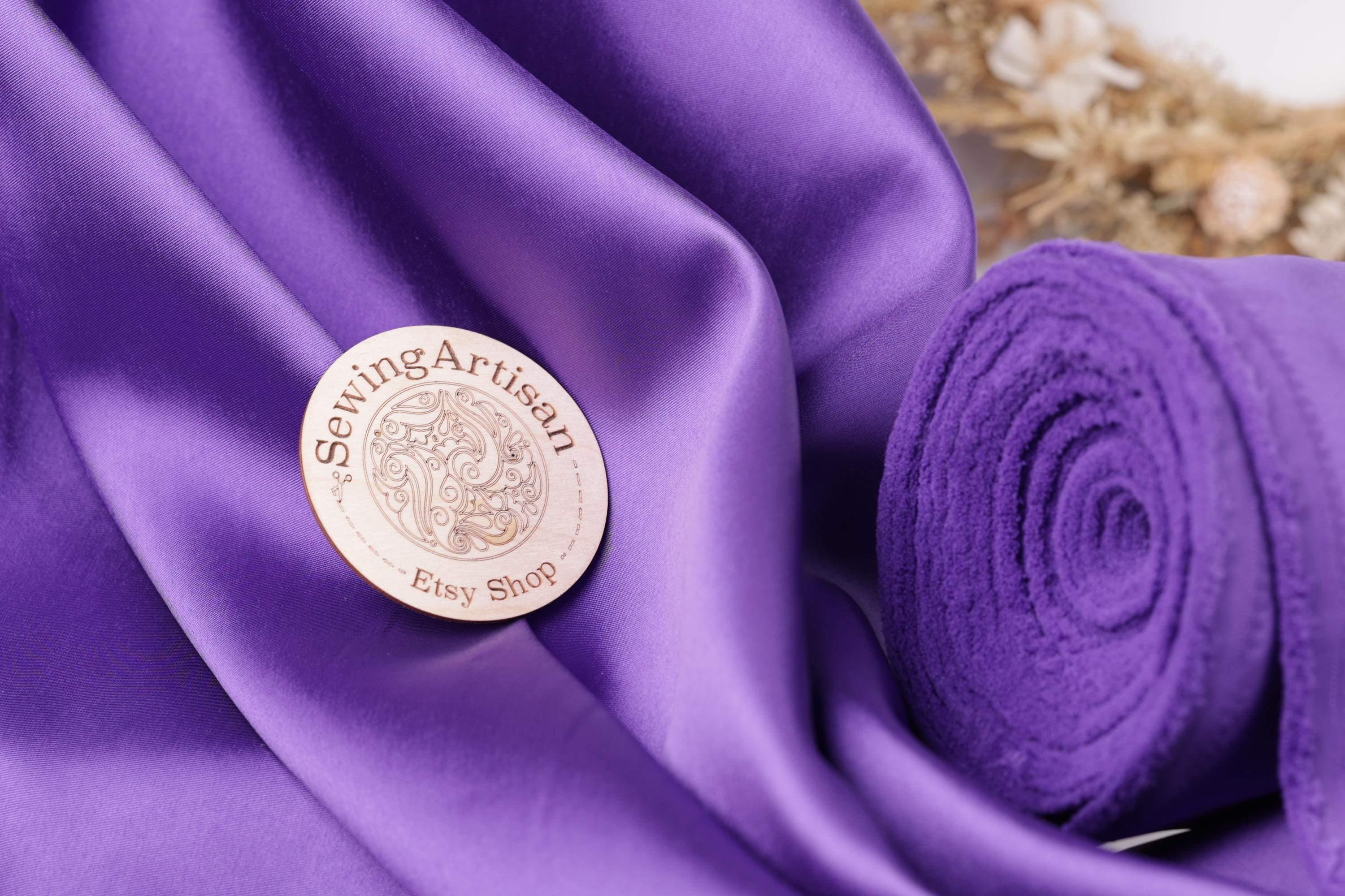 a close-up of a purple fabric with a wooden pin or badge prominently displayed on it, which reads "Sewing Artist" and "Ety Shop".