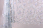 a close-up view of a delicate, light blue fabric with a repeating pattern of white and light purple flowers or leaves. The fabric appears to be made of sheer material, giving it a delicate and airy appearance. The pattern is intricate and detailed, with the flowers or leaves appearing to be hand-stitched or embroidered onto the fabric.