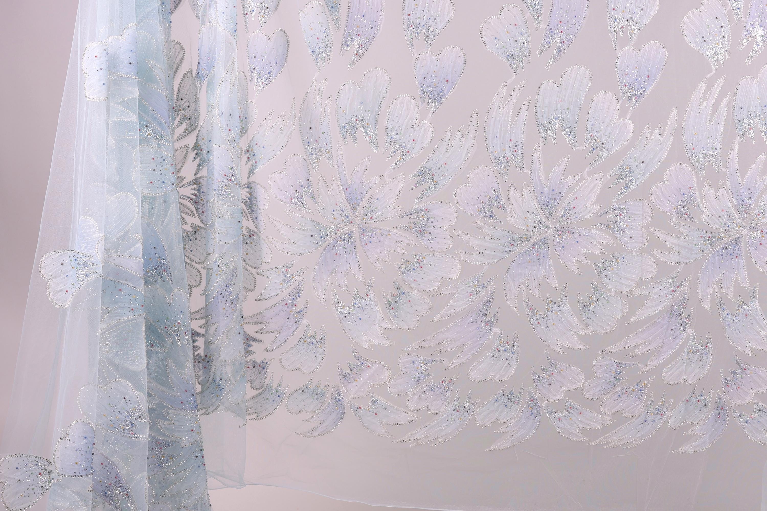 a close-up view of a delicate, light blue fabric with a repeating pattern of white and light purple flowers or leaves. The fabric appears to be made of sheer material, giving it a delicate and airy appearance. The pattern is intricate and detailed, with the flowers or leaves appearing to be hand-stitched or embroidered onto the fabric.