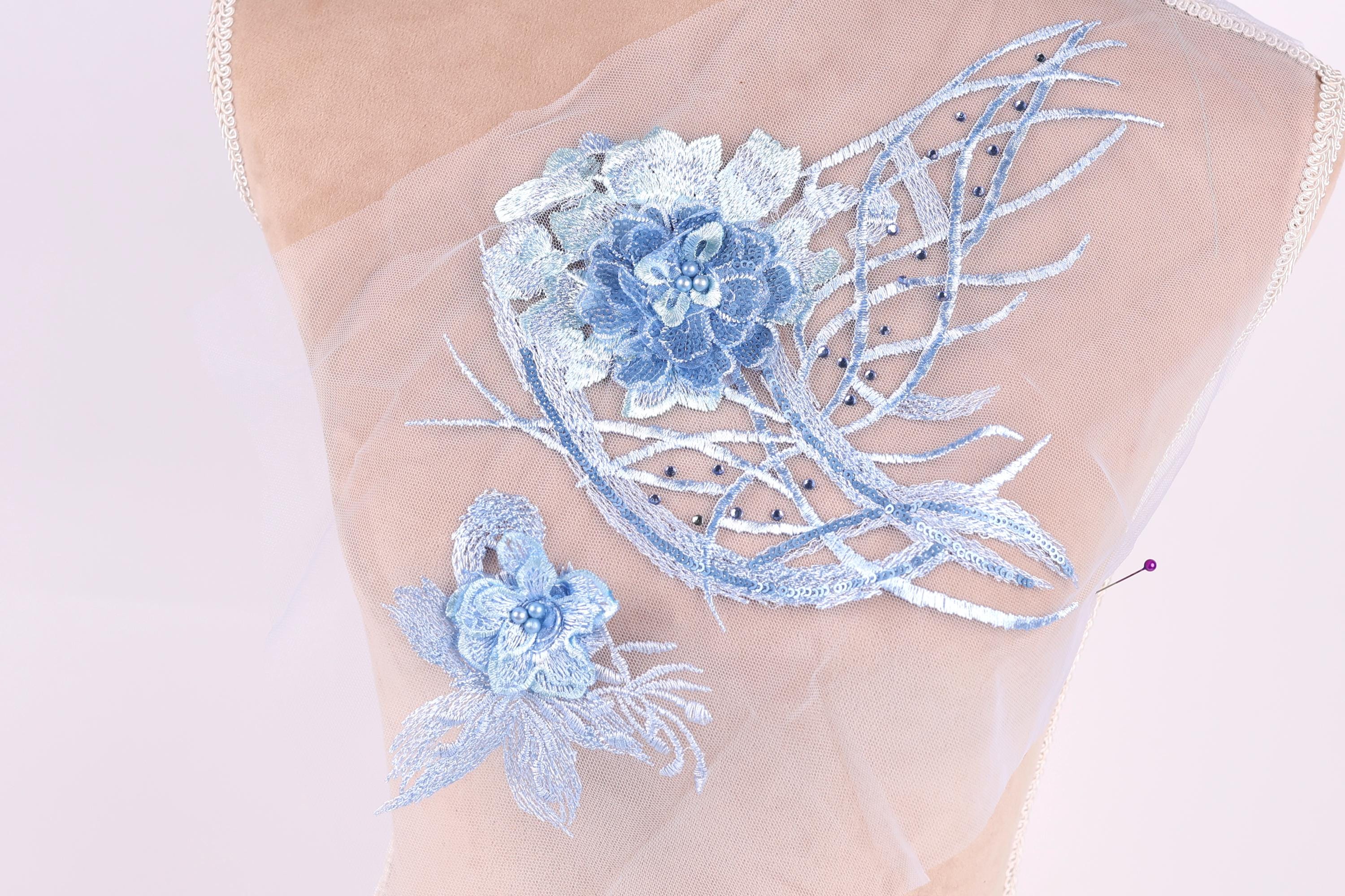a close-up view of a delicate, intricate embroidered design on a fabric surface. The design consists of two blue flowers, one larger and more prominent, and smaller blue flowers arranged in a floral pattern. The flowers are adorned with small blue beads, adding a touch of sparkle and detail to the overall design.