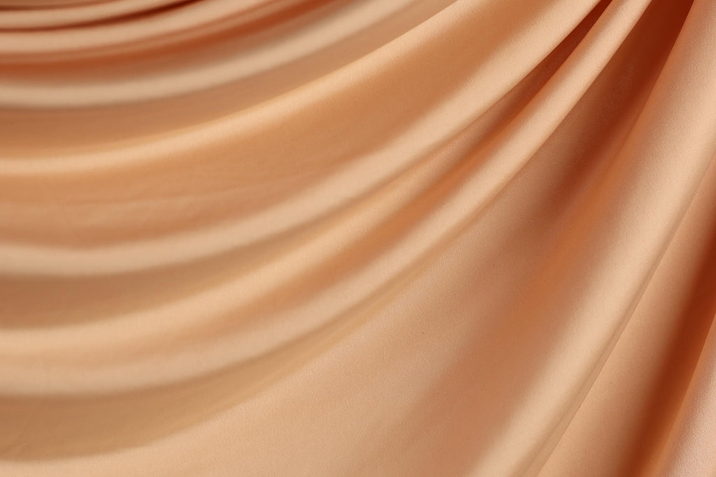 The image is a close-up of a flowing, crumpled fabric with a soft, peach-colored hue. The fabric appears to be made of a smooth, shiny material, possibly silk or satin, and has a slightly wrinkled texture. The image does not contain any discernible objects or actions, and the focus is solely on the fabric's texture and color.