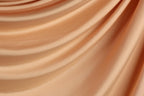 The image is a close-up of a flowing, crumpled fabric with a soft, peach-colored hue. The fabric appears to be made of a smooth, shiny material, possibly silk or satin, and has a slightly wrinkled texture. The image does not contain any discernible objects or actions, and the focus is solely on the fabric's texture and color.