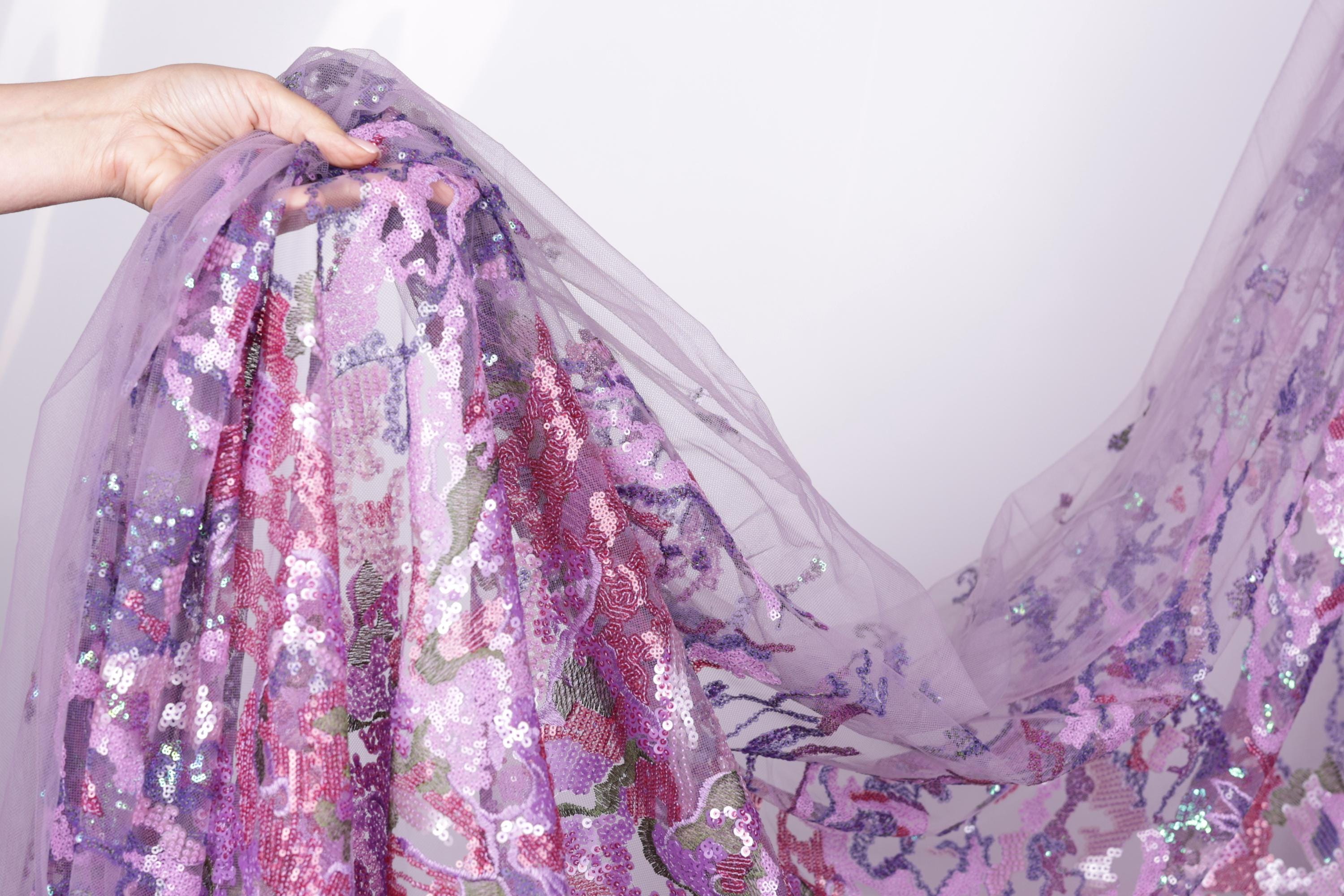 A hand is holding a piece of fabric with a floral pattern in shades of purple and pink.