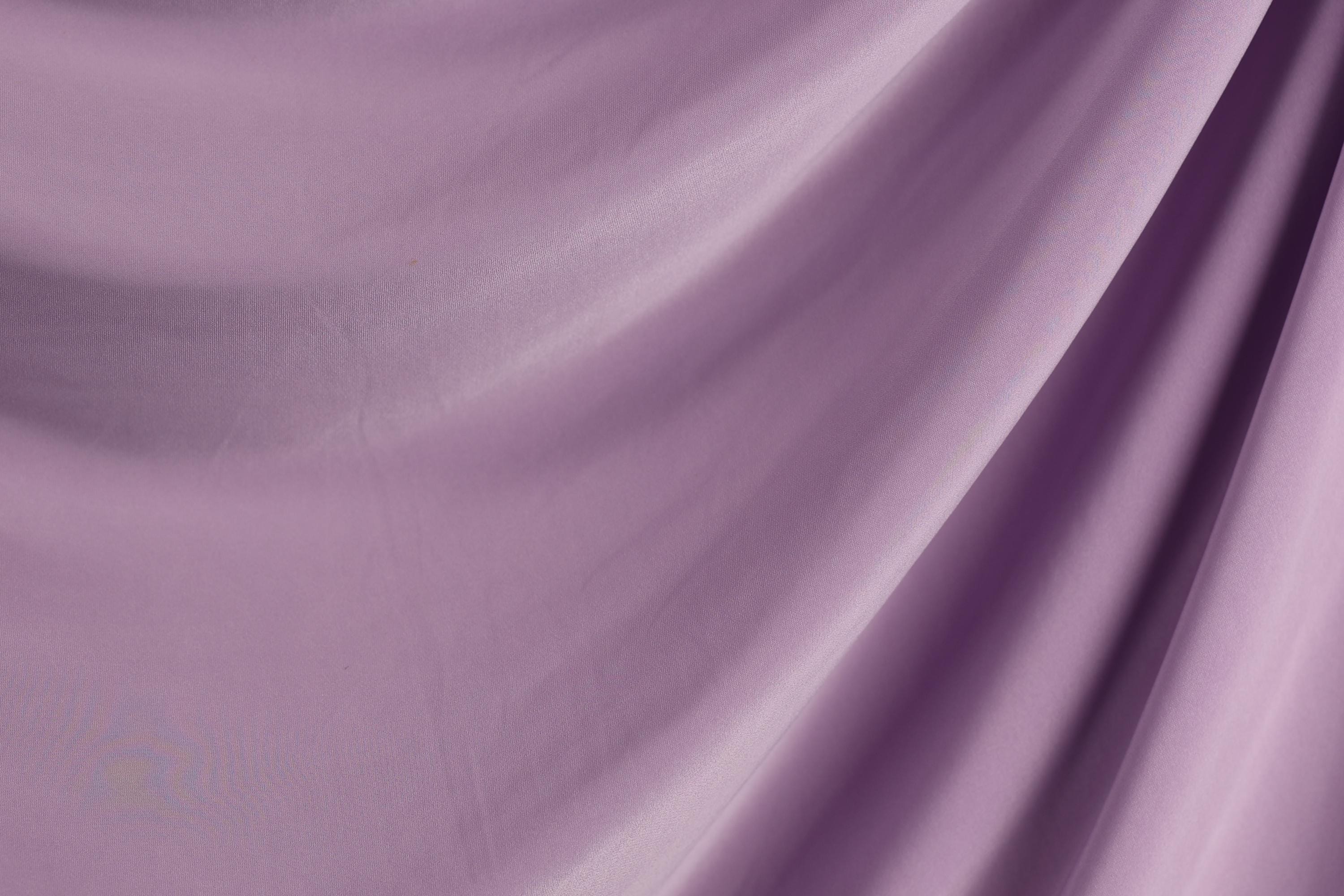a close-up view of a soft, flowing purple fabric, possibly a satin or chiffon material. The fabric appears to be made of silk or a similar luxurious material, with a smooth and shiny texture. The image does not contain any discernible objects or actions, and the focus is solely on the fabric's texture and color.