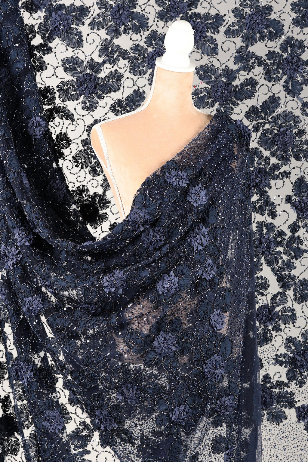 a mannequin wearing a black lace dress with a floral pattern. The dress is draped over the mannequin, creating a draped effect. The background is a white fabric with a black floral pattern.