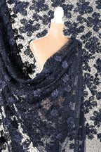 a mannequin wearing a black lace dress with a floral pattern. The dress is draped over the mannequin, creating a draped effect. The background is a white fabric with a black floral pattern.
