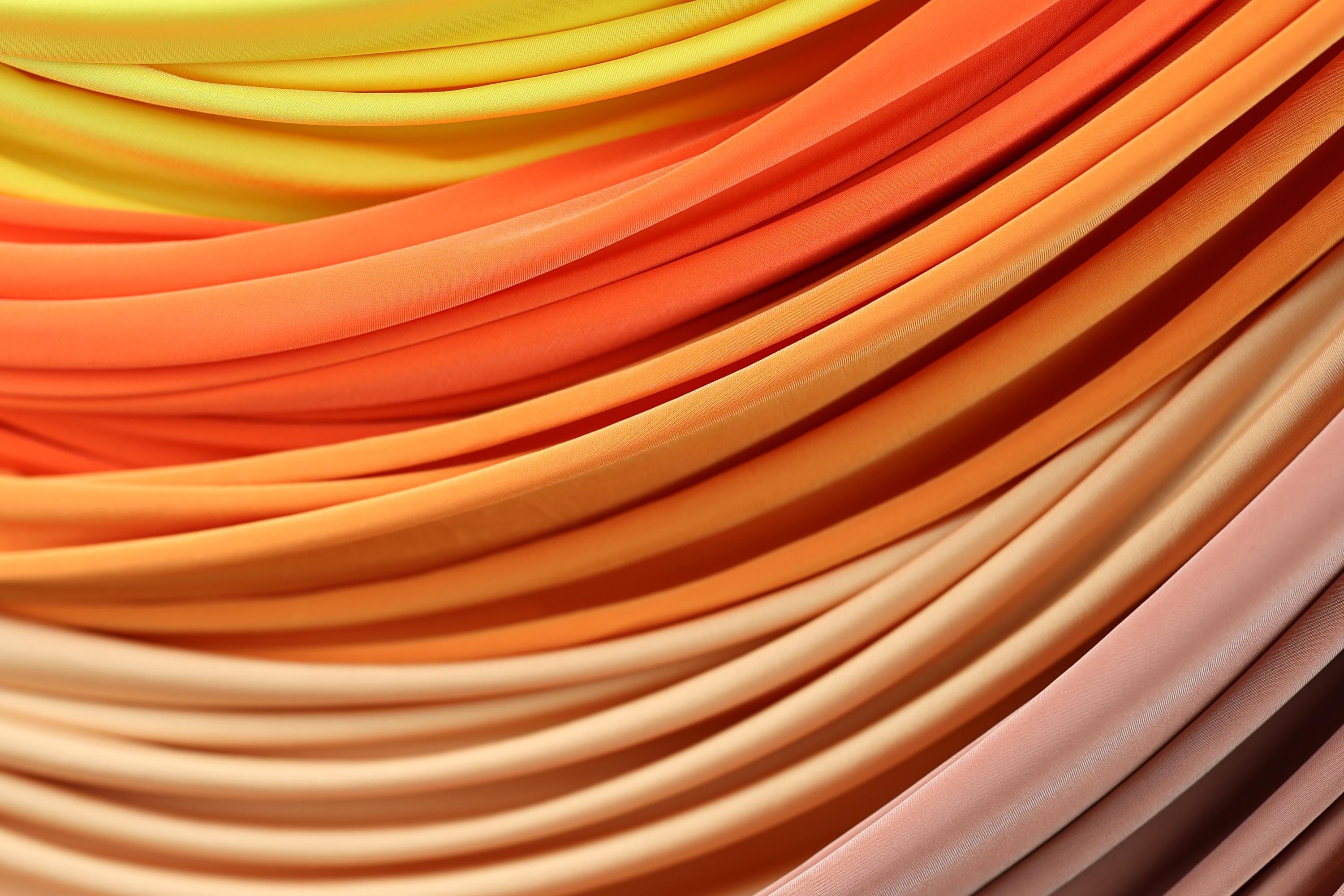 a close-up view of a vibrant, multi-colored fabric, with a gradient of warm hues ranging from yellow to orange to pink. The fabric appears to be folded or draped in a way that creates a visually striking and textured appearance.