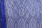 a close-up view of a fabric with a repeating pattern of blue sequin designs. The pattern consists of various diamond shapes and lines, creating a visually striking and intricate design. The fabric appears to be a piece of clothing or textile, with the blue sequins adding a vibrant and eye-catching element to the overall design.