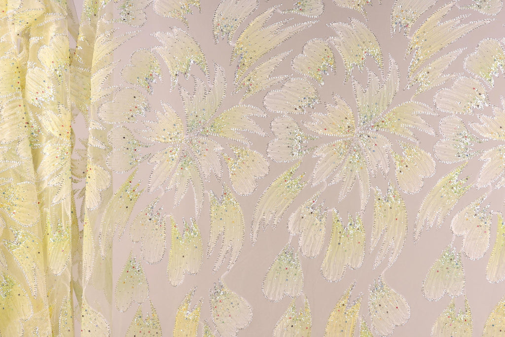 a close-up view of a fabric with a floral pattern, including yellow and white flowers and leaves. The fabric appears to be made of a shiny, glittery material, giving it a sparkly and eye-catching appearance.