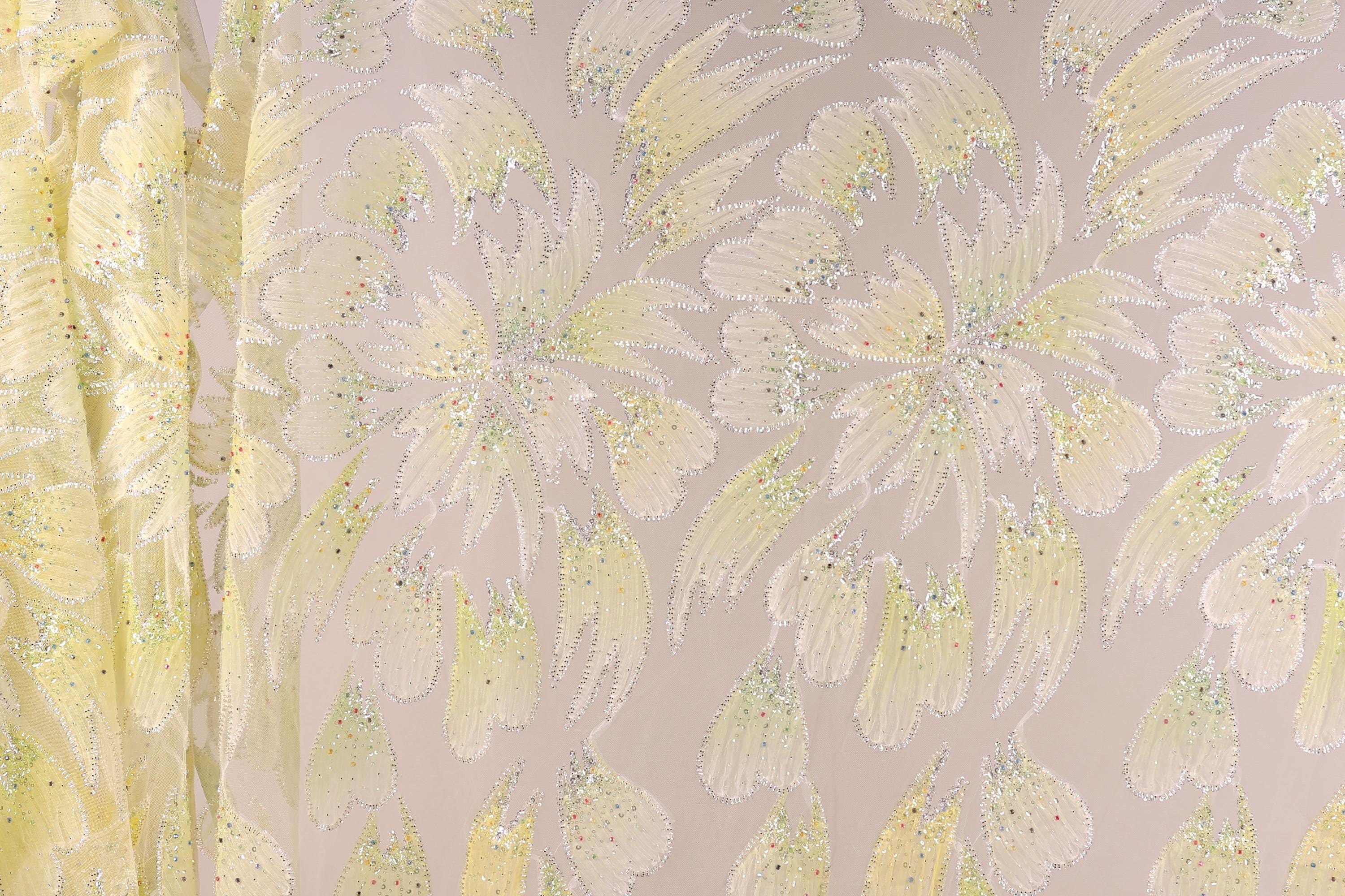 a close-up view of a fabric with a floral pattern, including yellow and white flowers and leaves. The fabric appears to be made of a shiny, glittery material, giving it a sparkly and eye-catching appearance.