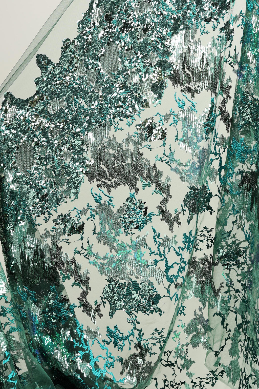 The image is a close-up view of a fabric with a pattern of green and silver sequins or sequin-like material. The fabric appears to be made of a shiny, metallic material, possibly metallic mesh or a similar type of material. The pattern on the fabric is intricate and detailed, with various shades of green and silver sequins or sequin-like elements creating a visually striking and textured appearance.