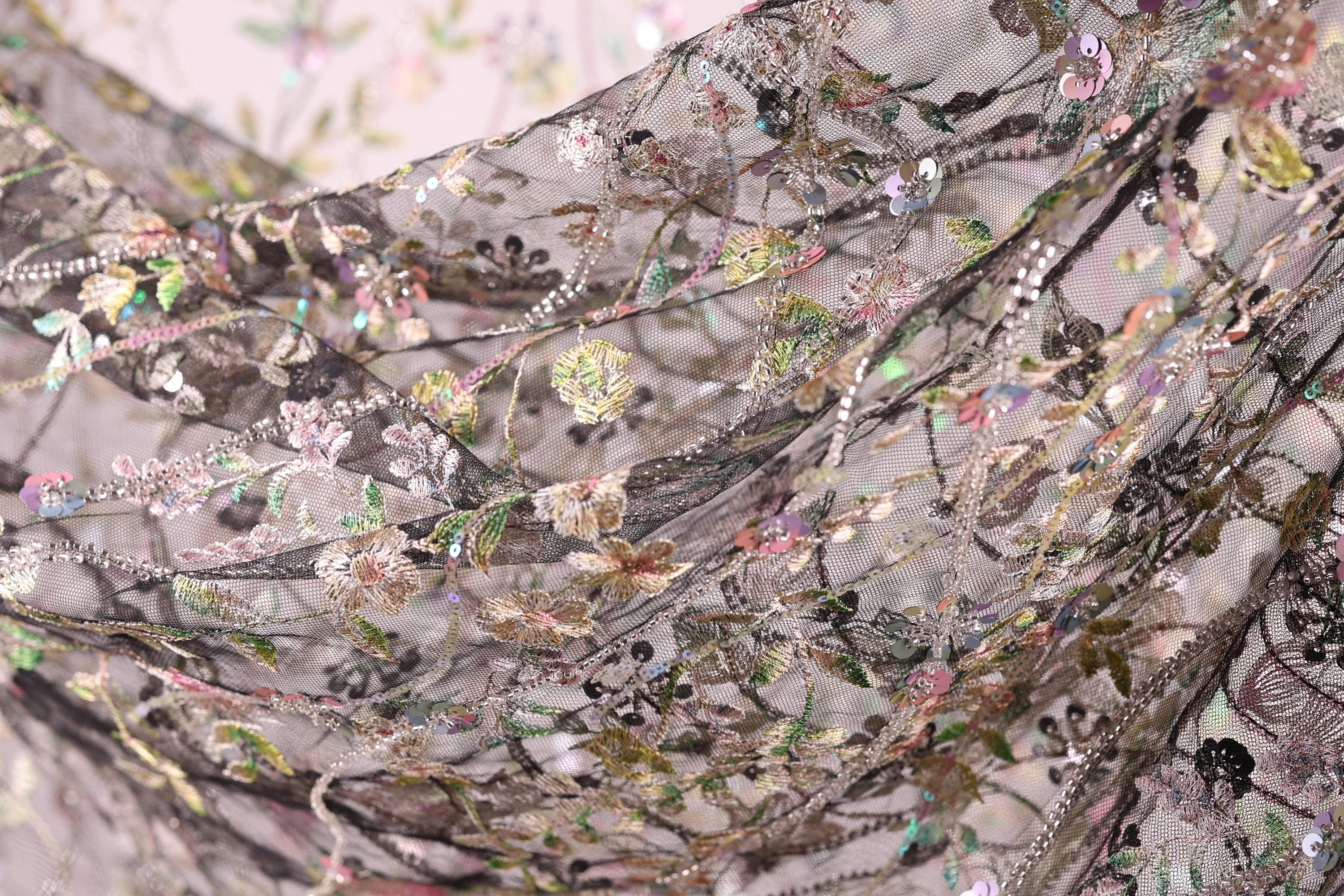The image is a close-up view of a delicate, intricately designed fabric with a floral pattern. The fabric appears to be made of a sheer material, with a variety of colors and textures, including shades of pink, green, and black. The intricate design and the presence of multiple flowers and leaves create a visually appealing and detailed appearance.