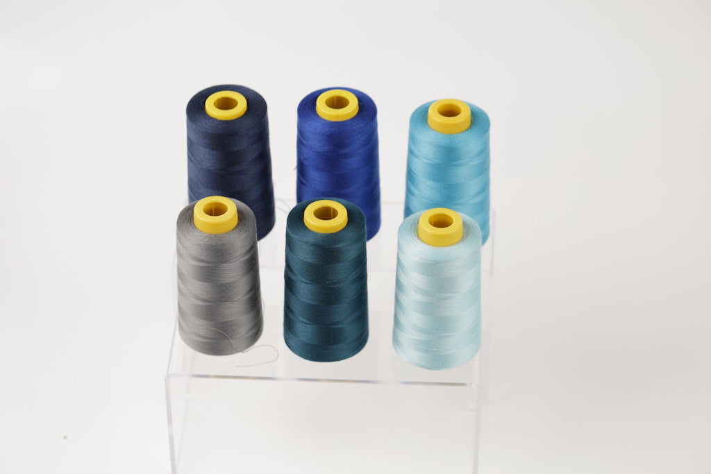 A collection of six spools of thread in various colors, including blue, gray, and light blue, arranged in a grid-like pattern on a clear stand.