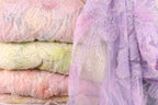 a close-up view of a stack of fabric swatches in various pastel colors, including pink, yellow, and purple. The fabric appears to be made of a sheer, lace-like material, with intricate beading and embroidery visible on the surface.