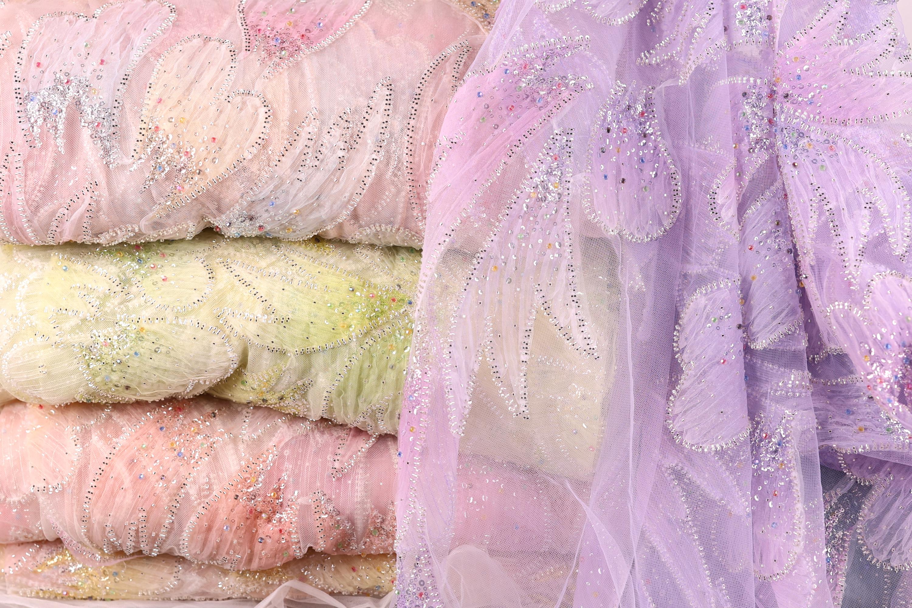 a close-up view of a stack of fabric swatches in various pastel colors, including pink, yellow, and purple. The fabric appears to be made of a sheer, lace-like material, with intricate beading and embroidery visible on the surface.