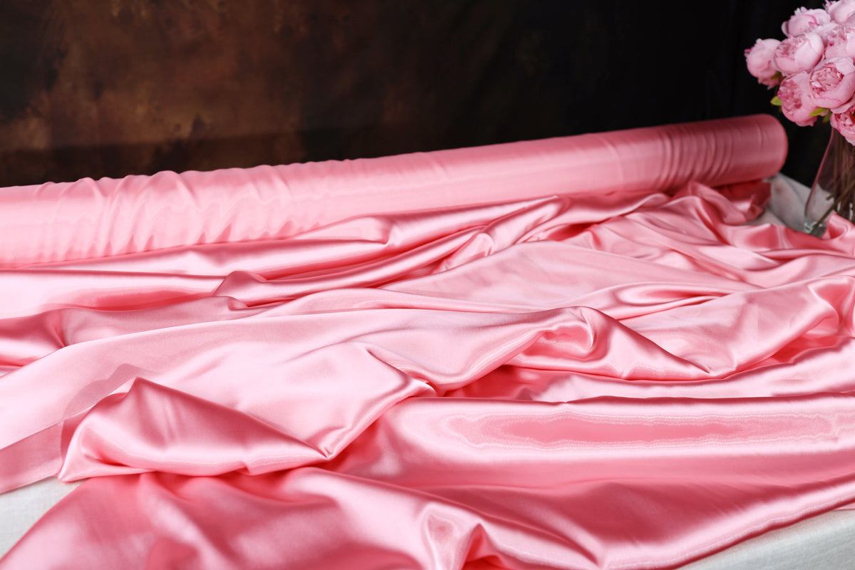 A close-up view of a pink satin fabric, with a vase of pink flowers visible in the background.