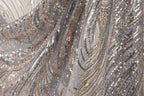 The image is a close-up of a fabric with a shimmering, sequined pattern in shades of silver, gold, and black. The fabric appears to be made of sheer material, giving it a delicate and elegant appearance.