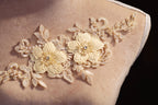 A close-up image of a delicate, beige lace flower embroidery on a fabric, with intricate details and a floral pattern.