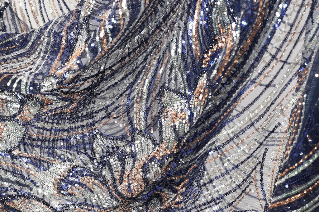 The image is a close-up view of a fabric with a complex, intricate pattern of sequins and metallic threads in shades of blue, silver, and gold. The fabric appears to be a piece of clothing or textile, with the intricate design and shimmering appearance suggesting a high level of craftsmanship and attention to detail.