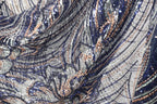 The image is a close-up view of a fabric with a complex, intricate pattern of sequins and metallic threads in shades of blue, silver, and gold. The fabric appears to be a piece of clothing or textile, with the intricate design and shimmering appearance suggesting a high level of craftsmanship and attention to detail.