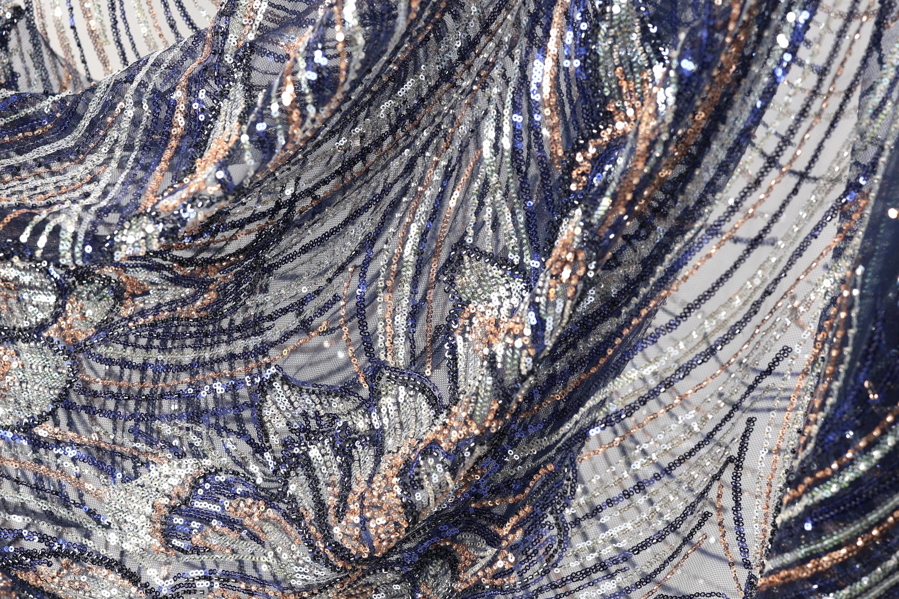 The image is a close-up view of a fabric with a complex, intricate pattern of sequins and metallic threads in shades of blue, silver, and gold. The fabric appears to be a piece of clothing or textile, with the intricate design and shimmering appearance suggesting a high level of craftsmanship and attention to detail.