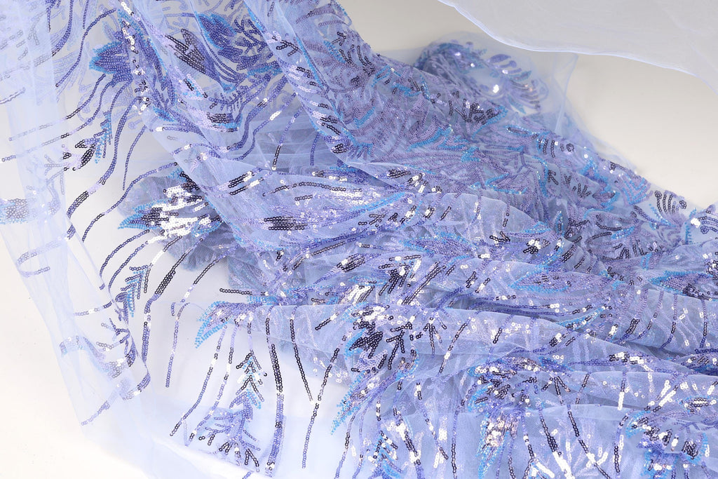 a close-up view of a fabric with a blue and purple floral pattern, featuring sequins and beads. The fabric appears to be a sheer material, with a delicate and intricate design.