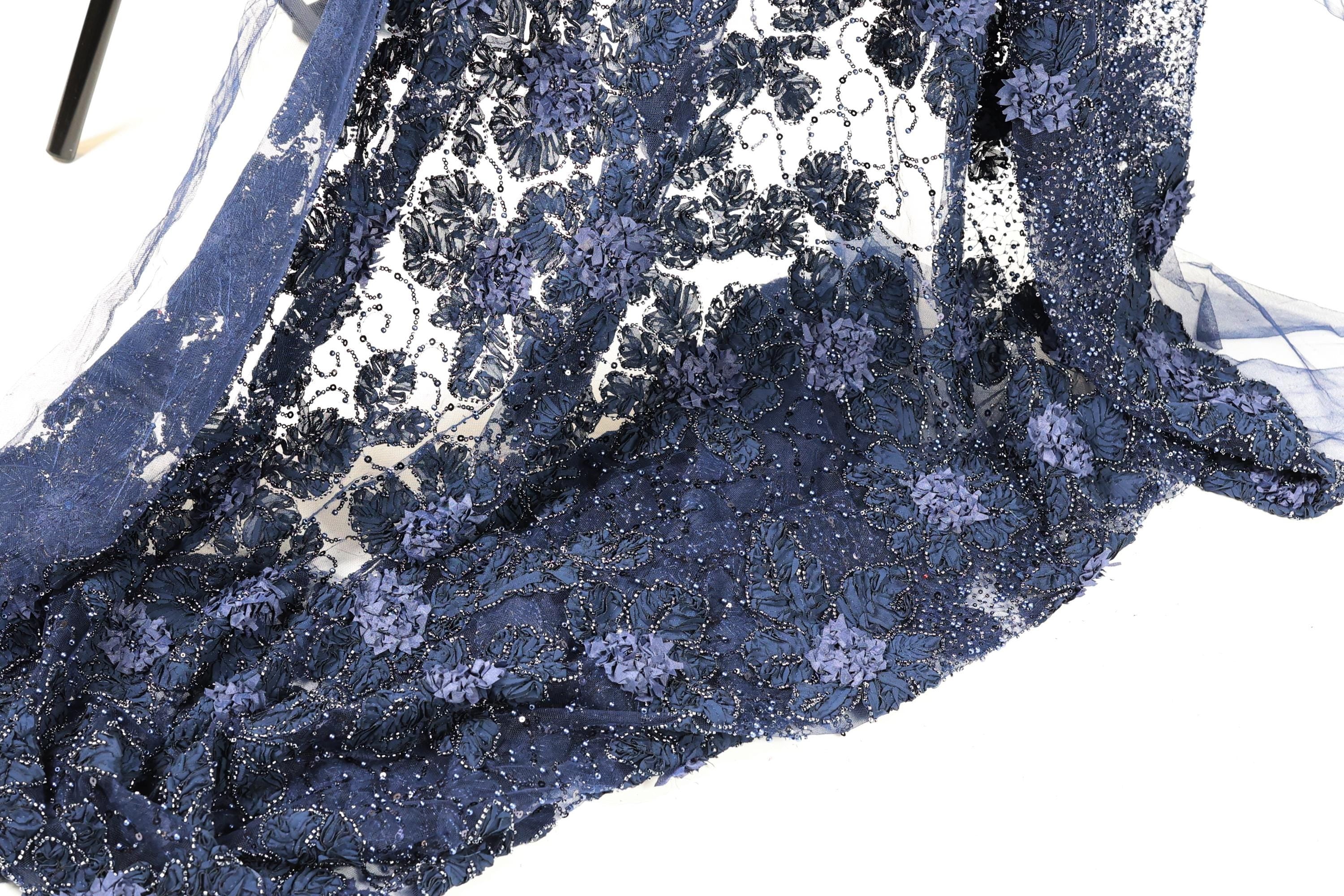 a close-up view of a fabric with a floral pattern, featuring blue and purple colors. The fabric appears to be made of lace or a similar material, with a delicate and intricate design.