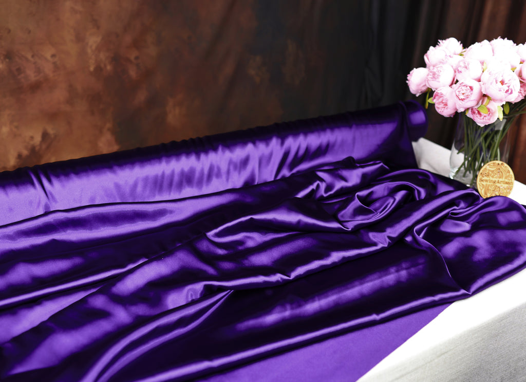 a purple satin fabric draped over a white surface, with a vase of pink flowers placed next to it.