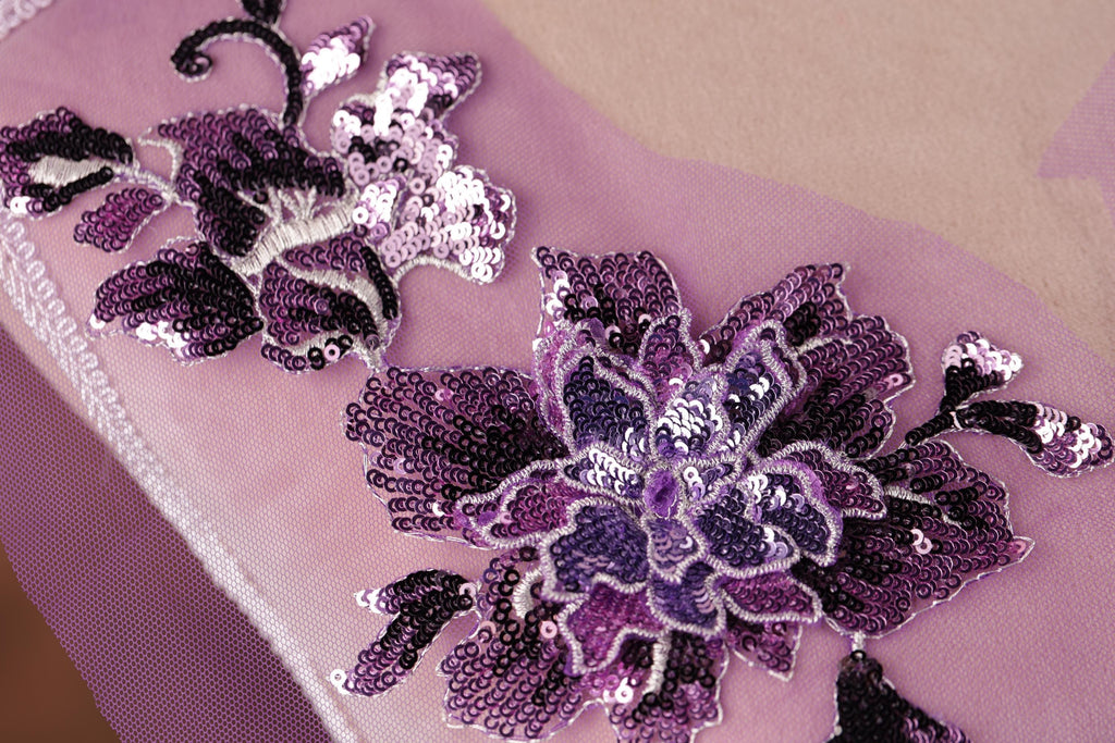 a close-up view of a delicate, intricately designed purple flower embroidered on a pink fabric.