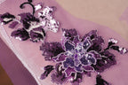 a close-up view of a delicate, intricately designed purple flower embroidered on a pink fabric.