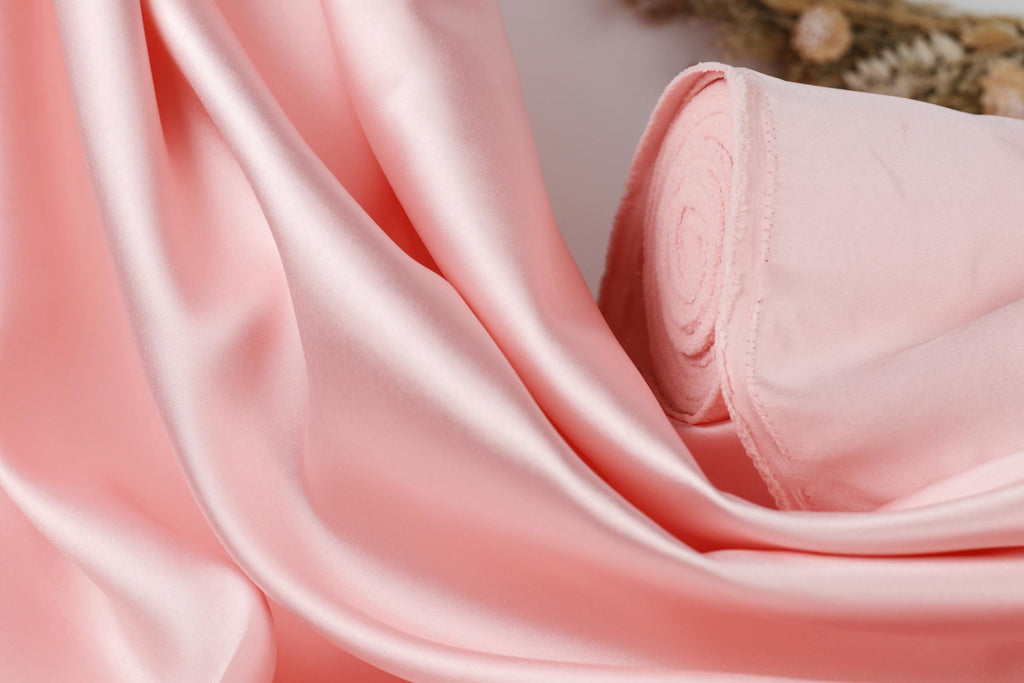 A close-up view of a soft, flowing pink satin fabric, with a blurred background that appears to be a plant or similar natural element.