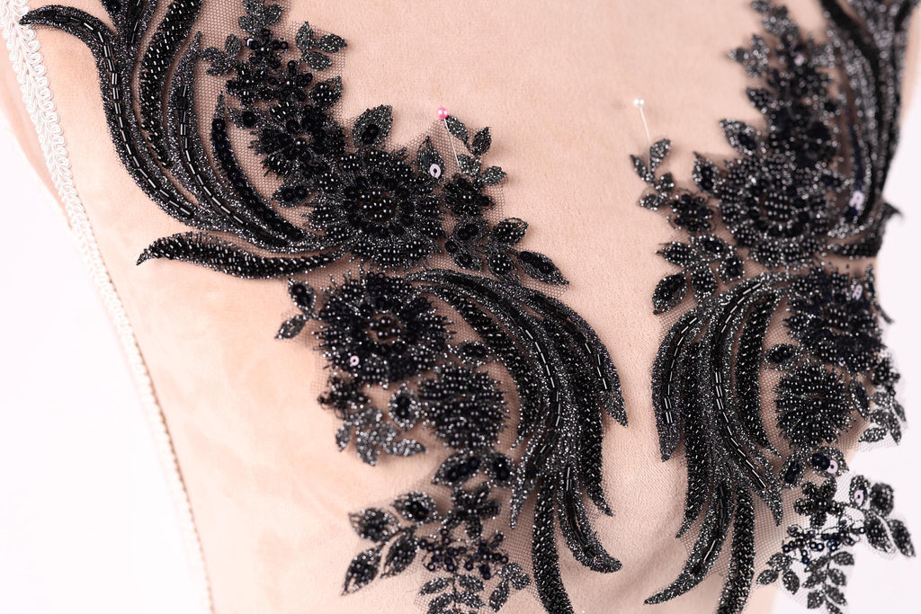 a close-up view of a delicate, intricate black lace design with floral and leaf-like patterns. The lace appears to be made of a shiny, black material and is embellished with small, round beads or sequins that add a touch of sparkle and elegance to the design.
