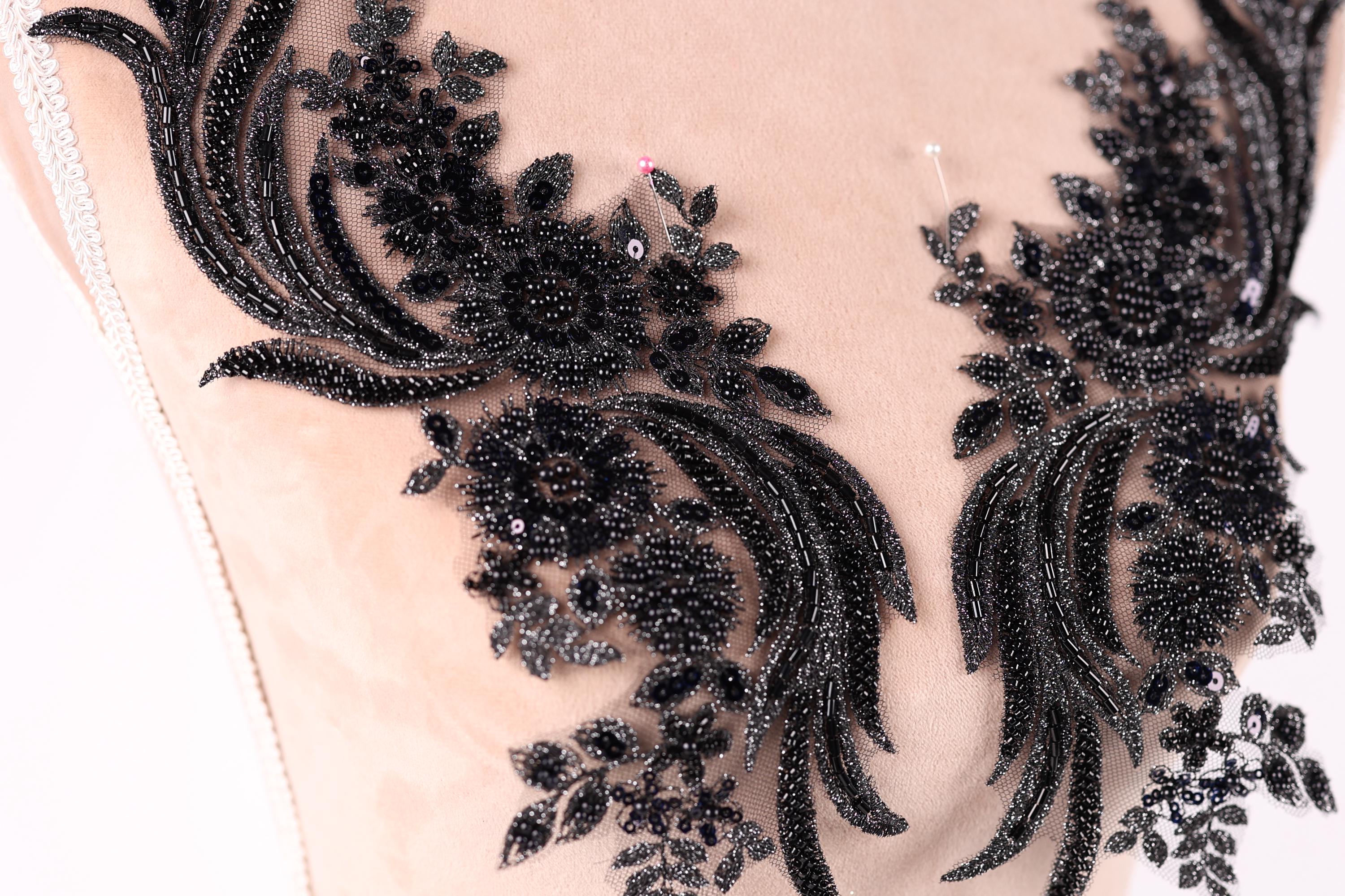 a close-up view of a delicate, intricate black lace design with floral and leaf-like patterns. The lace appears to be made of a shiny, black material and is embellished with small, round beads or sequins that add a touch of sparkle and elegance to the design.