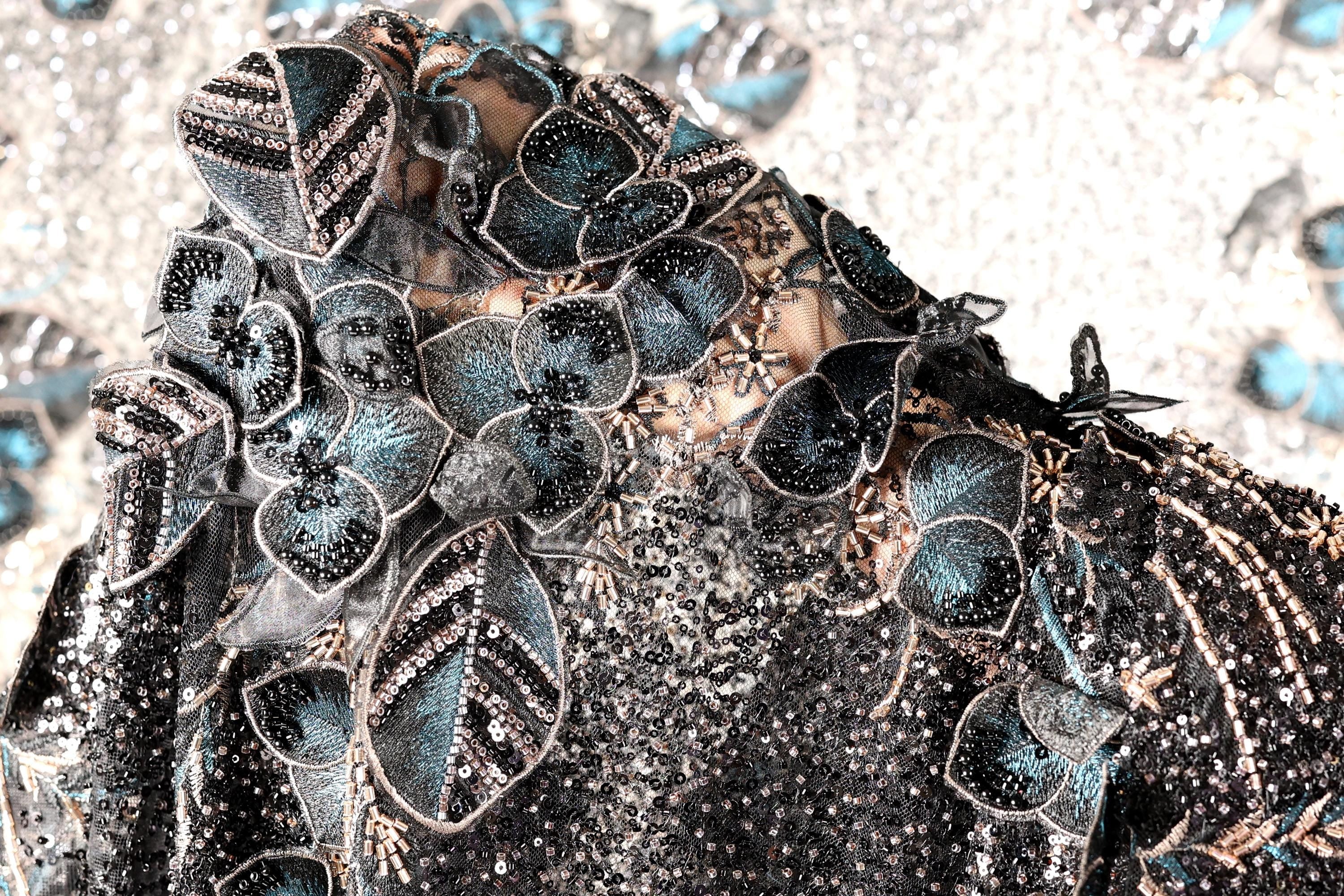 The image is a close-up of a black and blue sequined dress with intricate floral and leaf designs.